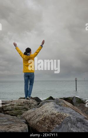 Rückansicht einer Frau, die an einem grau bewölkten Tag auf Felsen mit erhobenen Armen steht und auf das Meer blickt. Stockfoto