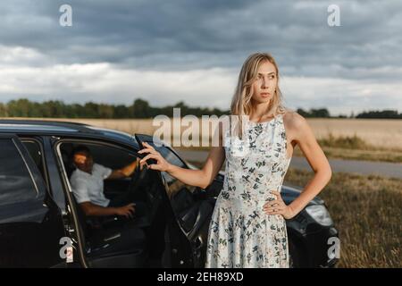 Blonde Frau, die in der Nähe der Autotür steht, und ein Mann, der im Auto sitzt. Paar im Auto Stockfoto