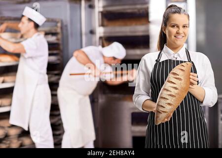 Junge Frau trägt eine Schürze und hält einen Laib Braunes Brot und Männer, die in einer Bäckerei arbeiten Stockfoto