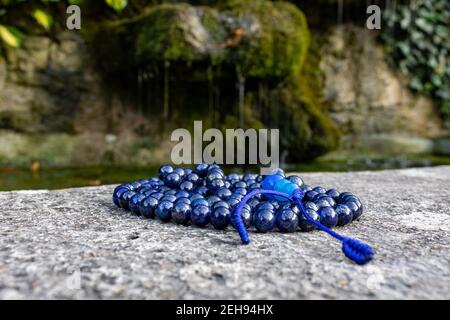 Blaue mala Perlen (Lapislazuli) auf Stein mit fließendem Wasser und grünen Blättern im Hintergrund. Achtsamkeit und Meditationszubehör. Stockfoto