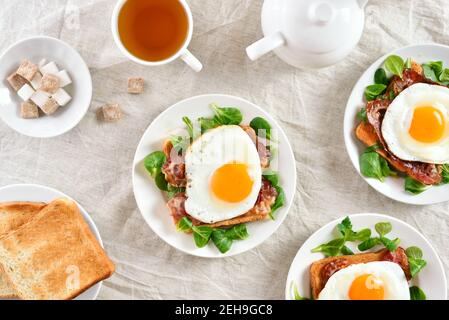 Offene Sandwiches mit Spiegeleiern, Speck und Gemüseblättern auf dem Teller auf hellem Hintergrund. Draufsicht, flach liegend Stockfoto