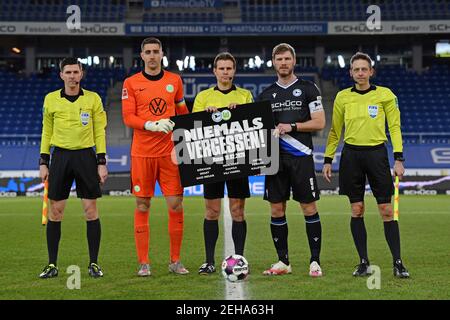 Bielefeld, Deutschland. Februar 2021, 19th. Fußball: Bundesliga, Arminia Bielefeld - VfL Wolfsburg, Matchday 22 in der Schüco Arena. Stefan Lupp (l-r), Schiedsrichterassistent, Koen Casteels, Mannschaftskapitän des VfL Wolfsburg, Felix Brych, Schiedsrichter, Fabian Klos, Mannschaftskapitän der Arminia Bielefeld, und Mark Borsch, Schiedsrichterassistent, halten ein Banner zum Gedenken an den rassistischen Angriff in Hanau vor einem Jahr. Ein Rechtsextremist hatte am 19. Februar 2020 in Hanau aus rassistischen Motiven neun Menschen erschossen. Anschließend hatte der Mann seine Mutter und dann sich selbst erschossen. Der Akt verursachte weltweiten Horror. Kredit: H/dpa/Alamy Live Nachrichten Stockfoto