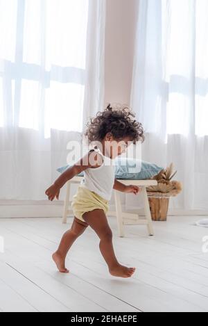 Kleine lockige afroamerikanische Mädchen spielen, Spaß haben, laufen um den hellen weißen Raum in einem minimalistischen Interieur im skandinavischen Stil. Stockfoto