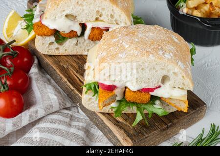 Burger mit Fischstäbchen frischer Salat, Tomate und Tartar-Sauce Set, auf Holzschneidebrett, auf weißem Hintergrund Stockfoto