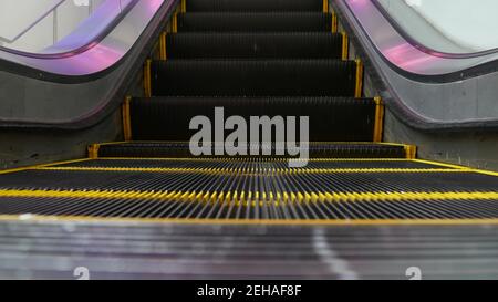 Blick auf die moderne Rolltreppe mit Looped-Perspektive. Automatisierter Aufzugsmechanismus. Gelbe Linie auf Treppe mit violettem Licht beleuchtet. Fut Stockfoto