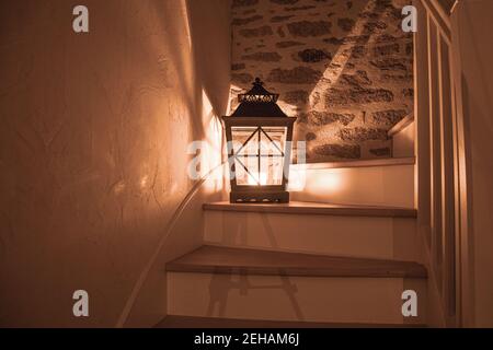 Eine große Laterne mit brennender Kerze auf einer Holztreppe und Schatten- und Lichtaspekten, die in alle Richtungen fallen, im Hintergrund eine Steinmauer Stockfoto