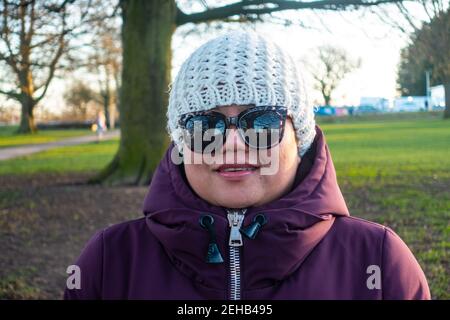 Porträt einer asiatischen Dame in einem Park mit warmem Wintermantel, Hut und Sonnenbrille im späten Nachmittagssonne an einem Tag im Februar 2021. Stockfoto