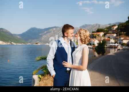 Braut und Bräutigam umarmen sich am Pier in Perast, der Bräutigam hält seine Hand in der Tasche Stockfoto