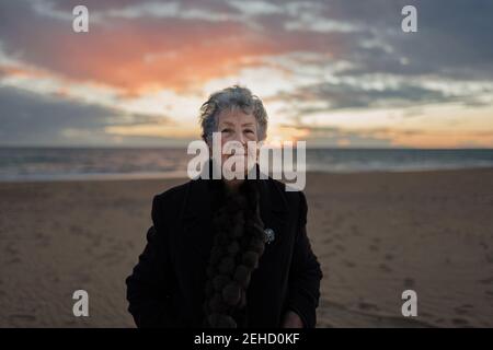 Ältere Frau in warmer legerer Kleidung, die den Sonnenuntergang über dem Meer bewundert Während der Ruhe allein am Sandstrand Stockfoto