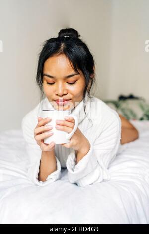 Lange Haare brünett asiatische Frau auf dem Bett zu Hause mit einer Tasse Kaffee liegen Stockfoto