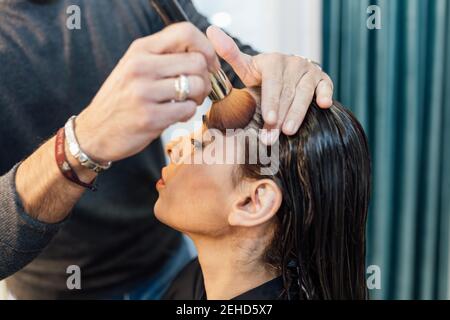 Crop unkenntlich männlichen Visagiste mit Pinsel beim Auftragen von Pulver auf Gesicht des selbstbewussten ethnischen weiblichen Modells Stockfoto