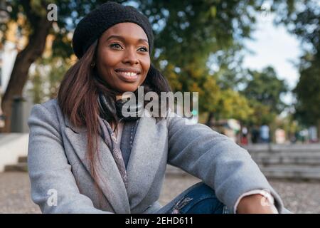 Sorglose afroamerikanische Frau in Oberbekleidung sitzen auf der Straße, während Wegschauen in Kontemplation Stockfoto