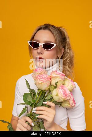 Junge schöne Frau in weißem Outfit und trendige Sonnenbrille halten Zarte rosa Rosen, während auf gelbem Hintergrund im Foto stehen studio Stockfoto