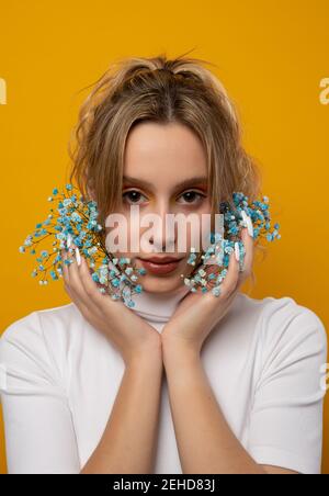 Attraktive junge weibliche stehend mit blauen zarten Gypsophila Blüten auf Das Gesicht auf gelbem Hintergrund im Studio Blick auf die Kamera Stockfoto