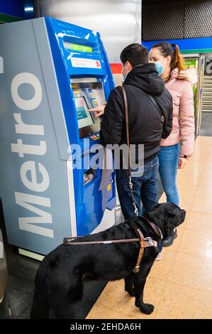 Ganzer Körper des Weibchens in der Maske hilft dem Menschen mit Blindheit Stehen mit Blindenhund beim Kauf von U-Bahn-Ticket an Verkaufsautomaten Maschine Stockfoto