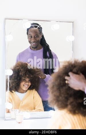 Crop African American Vater Kämmen lockiges Haar des Mädchens sitzen In der Nähe des Spiegels und lächelnd sanft Stockfoto