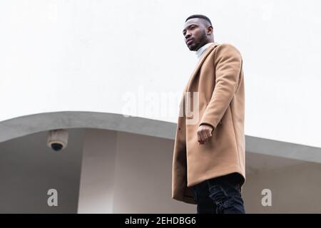 Von unten Seitenansicht des unemotionalen jungen verträumten Afroamerikaners Männchen schaut unter weißem Himmel in der Stadt weg Stockfoto