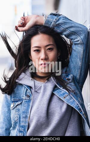 Asiatische Brünette Frau mit langen Haaren an der Wand gelehnt und Blick auf die Kamera Stockfoto