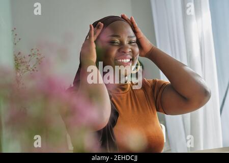 Glückliche junge stilvolle afrikanische Dame in traditionellen Hijab mit Händen Auf dem Kopf und Blick in Spiegel in Sonnenlicht zu Hause Stockfoto