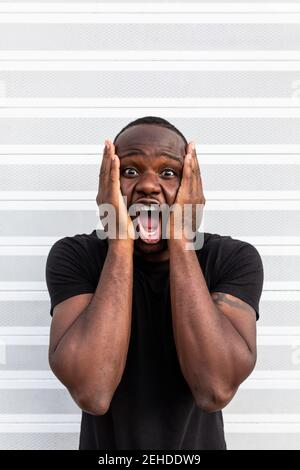 Junge erstaunt afroamerikanischen Mann in schwarzem T-Shirt schreien Beim Berühren des Gesichts und beim Betrachen der Kamera auf hellem Hintergrund Stockfoto
