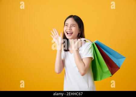 Junge lächelnde asiatische Frau hält mehrfarbige Einkaufstaschen und kündigt mit den Händen zum Mund auf hellgelben Hintergrund. Stockfoto