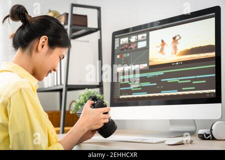 Asiatische Frau hält eine Kamera und mit Anwendung Video-Editor arbeitet auf dem Computer mit Filmmaterial auf Holztisch. Stockfoto