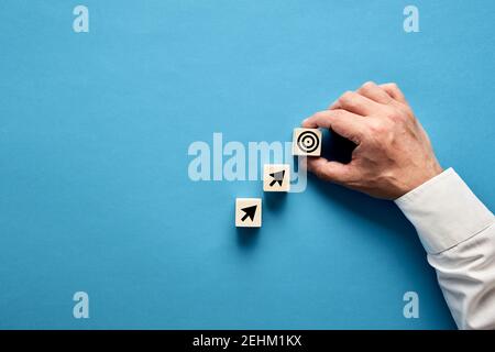 Pfeil und Zielsymbole auf Holzblöcken mit einer Geschäftsmann-Hand, die das Zielsymbol platziert. Ziel- oder Agendaeinstellung im Geschäftskonzept. Stockfoto