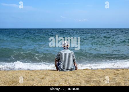 Mann in gestreiftem Hemd und panama-Hüte sitzt am Meer und entspannt, schaut in grenzenlose blaue Distanzierung. Urlaub, Strandurlaub. Stockfoto