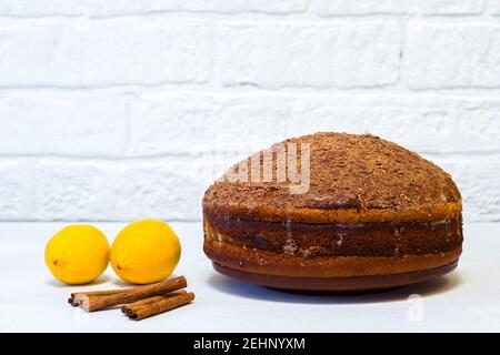 Zitronenkuchen bestreut mit geriebener Schokolade mit Zitronen und Zimt auf einem weißen Tisch an einer weiß verzierten Wand. Das Konzept der Herstellung von Dessert. Stockfoto