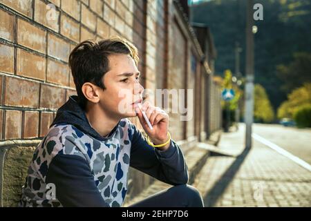 Rauchen. Portrait eines Teenagers, der mit einem nachdenklichen Blick eine Zigarette an den Mund hält. Backsteinmauer und Straße im Hintergrund. Das Konzept von n Stockfoto