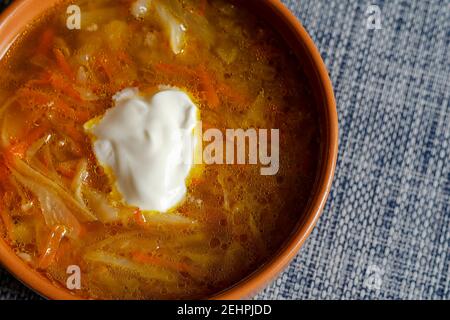 Kohlsuppe mit saurer Sahne in einer braunen irdenen Schüssel auf einer blauen Tischdecke. Eine Portion Fertigsuppe. Draufsicht aus einem Winkel, selektiver Fokus. Stockfoto