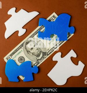 Der amerikanische fünf-Dollar-Schein ist unter den Puzzleteilen versteckt. Blau-weiße Puzzles mit Geld auf braunem Hintergrund. Draufsicht, flach liegend. Stockfoto