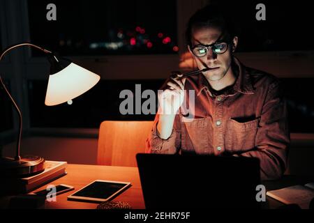 Kaukasische männliche Freiberufler arbeiten spät in der Nacht gestresst beißen Stift Bei der Suche nach Clients auf dem Laptop Stockfoto