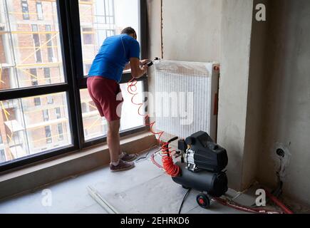 Rückansicht der Klempnerfüllrohre mit Druckluft zur Überprüfung auf Undichtigkeiten bei Neuinstallation des Heizsystems. Junger Mann mit Luftkompressor. Konzept der Renovierung und Gas Dichtheitsprüfung Stockfoto