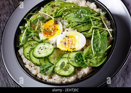 Quinoa-Schüssel mit Ei, Sonnenblumenkraut, Gurke, Avocado, Babyspinat in einer schwarzen Schüssel auf einem dunklen Holztisch, Nahaufnahme, türkische Küche Stockfoto