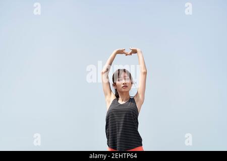 Junge asiatische Frau in Sportkleidung Stretching Arme im Freien gegen blau Himmel Stockfoto