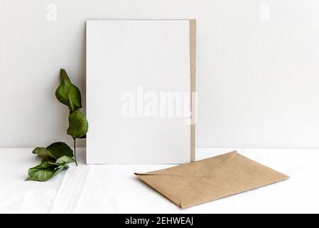 Modische Stock Schreibwaren Hintergrund - weiße Karte, Kraftpapier Umschlag und getrocknete Pflanzen und Blumen auf einem weißen Tisch. Hochzeit femininer Hintergrund. Leer Stockfoto