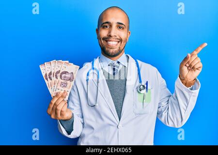 Hispanic erwachsenen Arzt Mann trägt medizinische Uniform hält 500 mexikanische Pesos lächelnd glücklich zeigt mit Hand und Finger auf die Seite Stockfoto