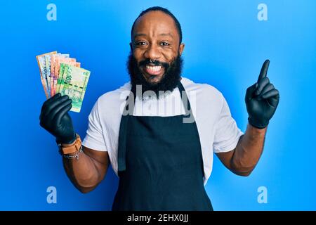 Junger afroamerikanischer Mann mit professioneller Schürze, der Südafrika hält rand Banknoten lächelnd glücklich zeigt mit Hand und Finger auf Die Seite Stockfoto