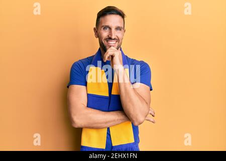Schöner Mann mit Bart Fußball Hooligan jubeln Spiel lächelnd Blick zuversichtlich auf die Kamera mit gekreuzten Armen und Hand am Kinn. Denken positiv. Stockfoto