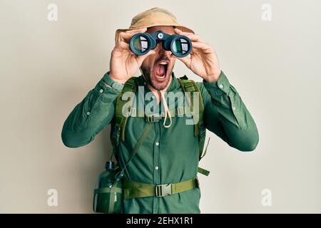 Schöner Mann mit Bart trägt Explorer Hut Blick durch Fernglas wütend und verrückt schreien frustriert und wütend, schreien vor Wut. Wut und Stockfoto