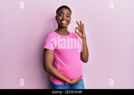 Junge afroamerikanische Frau erwartet ein Baby, berühren schwangeren Bauch lächelnd positive tun ok Zeichen mit Hand und Fingern. Erfolgreicher Ausdruck. Stockfoto