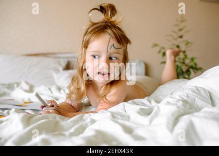 Mädchen zeichnet einen Regenbogen auf weißem Papier mit Filzstiften auf dem Bett. Kinder spielen am Morgen zu Hause. Schelmisch frech Baby, verschmierte Hände, Füße Stockfoto