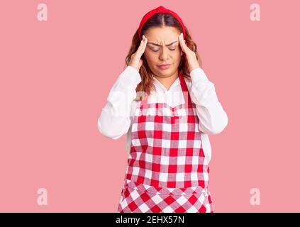Junge lateinische Frau trägt Schürze leiden unter Kopfschmerzen verzweifelt und gestresst, weil Schmerzen und Migräne. Hände auf dem Kopf. Stockfoto