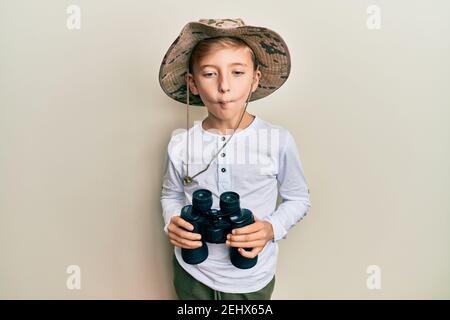 Kleiner kaukasischer Junge Kind trägt Explorer Hut halten Fernglas machen Fisch Gesicht mit Mund und blinzeln Augen, verrückt und komisch. Stockfoto