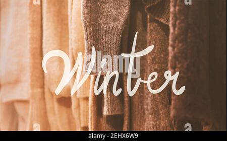 Auf Kleiderbügel in der Garderobe hängen beige und braun warme gemütliche Strickpullover, beleuchtet durch Licht, und auf der Oberseite ist eine weiße Inschrift "Winter gemalt Stockfoto