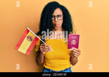 Mittelalter afroamerikanische Frau hält spanien Flagge und Pass machen Fisch Gesicht mit Mund und blinzeln Augen, verrückt und komisch. Stockfoto