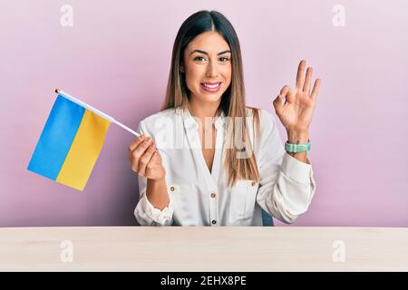 Junge hispanische Frau hält ukraine Flagge auf dem Tisch sitzen ok Zeichen mit den Fingern, lächelnd freundlich gestikulieren ausgezeichnetes Symbol Stockfoto