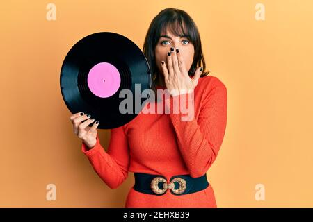 Junge hispanische Frau hält Vinyl-Disc über den Mund mit der Hand, schockiert und Angst vor Fehler. Überrascht Ausdruck Stockfoto