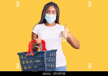 Junge afroamerikanische Frau mit Zöpfen tragen Warenkorb und medizinische Maske lächelnd glücklich und positiv, Daumen nach oben tun ausgezeichnet und Zustimmung si Stockfoto